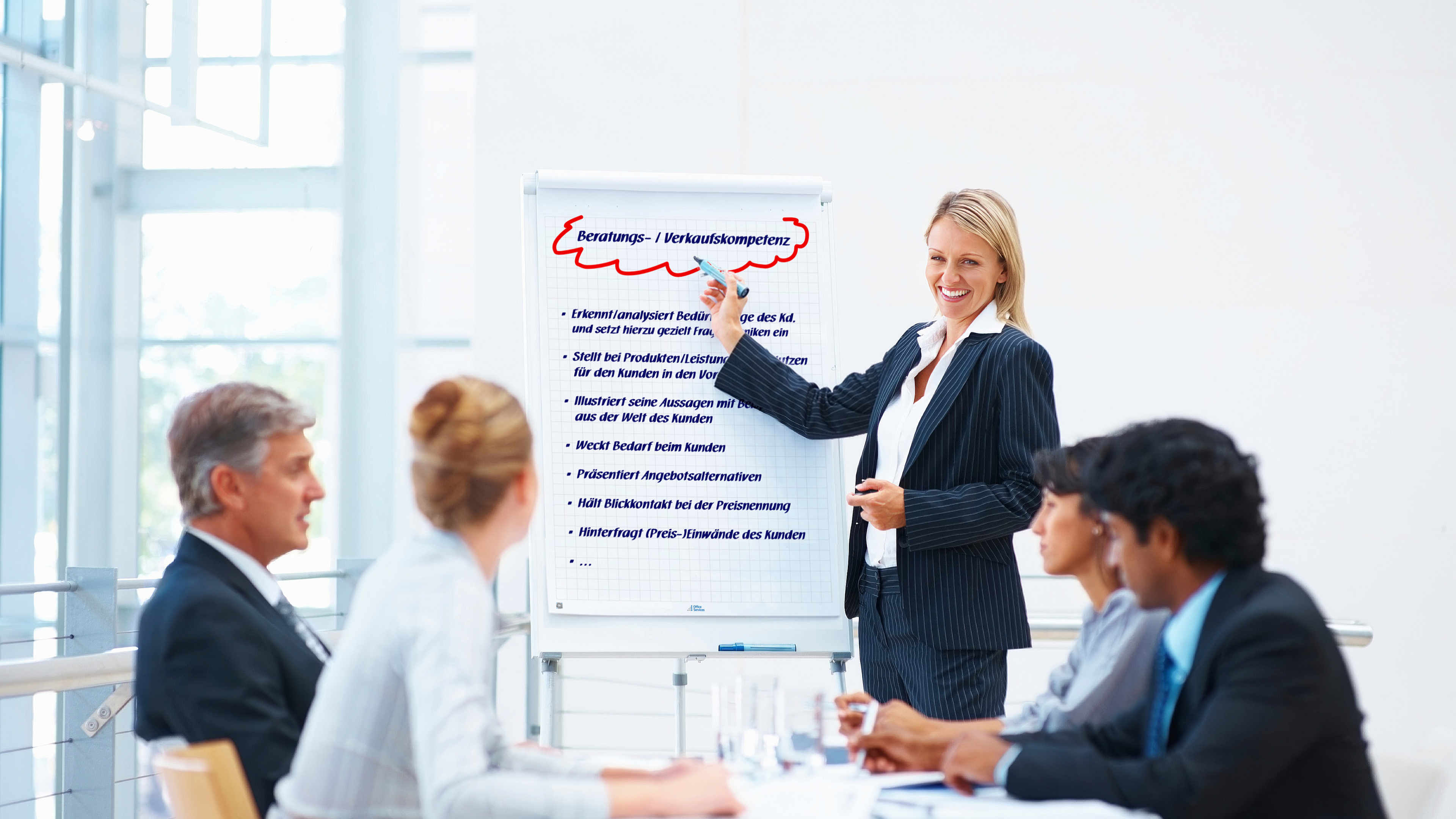 Eine Trainerin zeigt auf ein Flipchart während sie in Richtung der Teilnehmenden der Weiterbildung blickt.